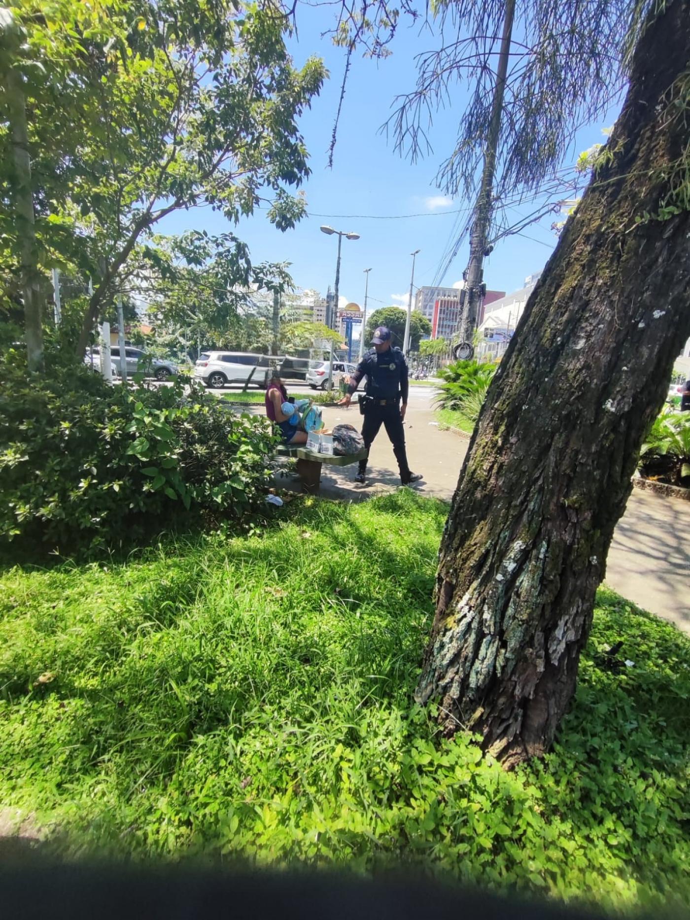 praça com gramado. ao fundo há uma mulher com um bebê no colo sentada em um banco. Um guarda municipal uniformizado está ao lado dela. #paratodosverem 