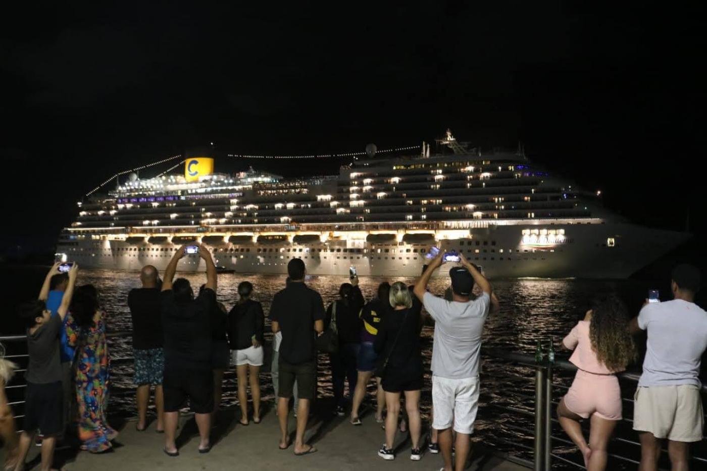 navio saindo à noite e público assistindo em terra. #paratodosverem
