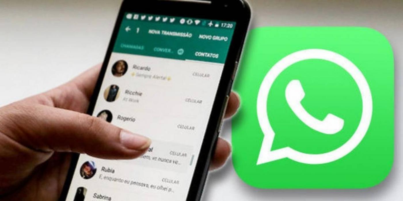 Imagem em close mostra uma mão segurando um smartphone aberto no aplicativo Whatsapp. Ao fundo, o logotipo do whatsapp. #Pracegover