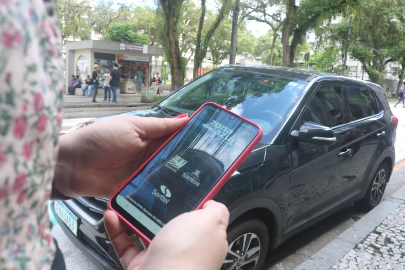 Mulher segura celular com aplicativo do Zona Azul e carro ao fundo. #pratodosverem