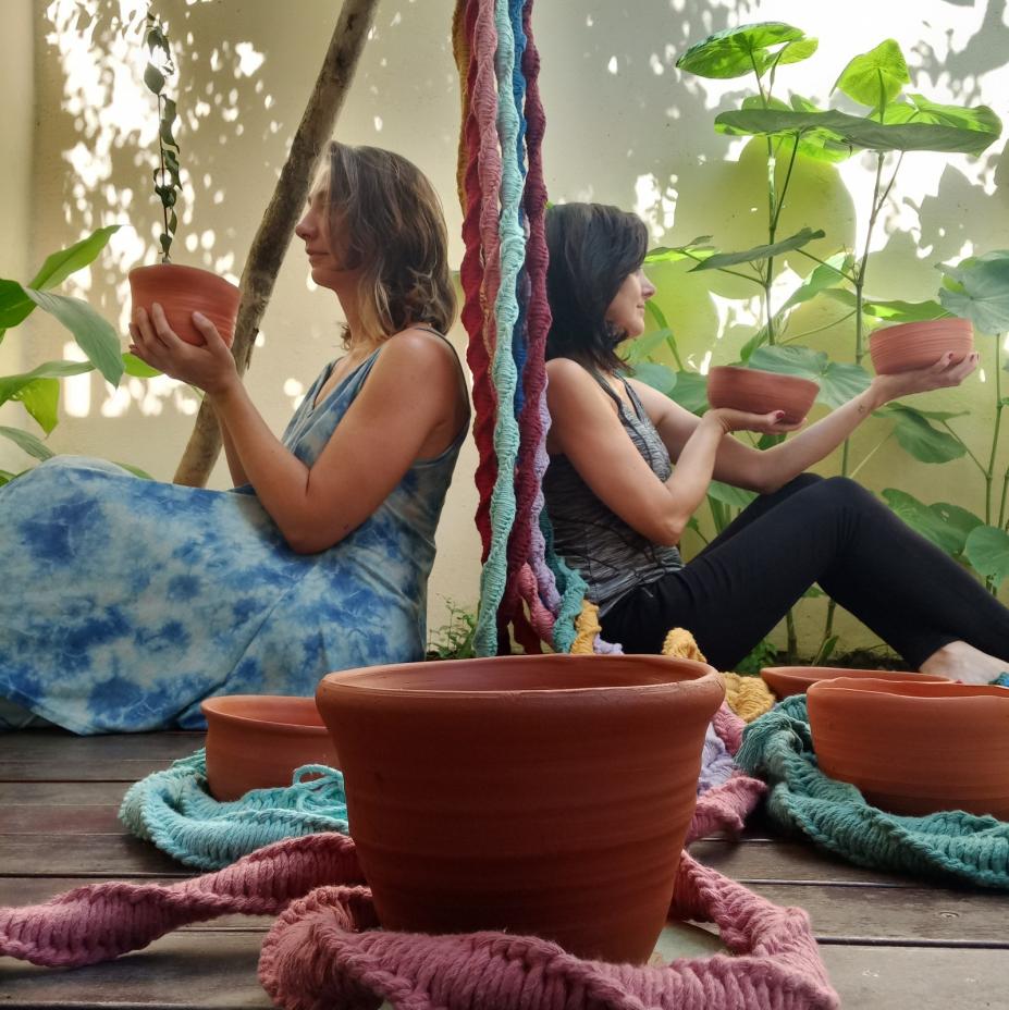 duas mulheres estão sentadas no chão, uma de costas para a outra. Ambas seguram vasos. Elas estão em meio a cordas de macramê. #paratodosverem 