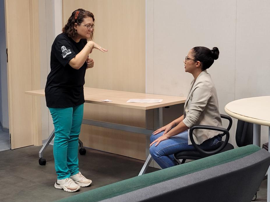 Mulher em pé faz gesto em Libras para outra mulher que está sentada. #paratodosverem 