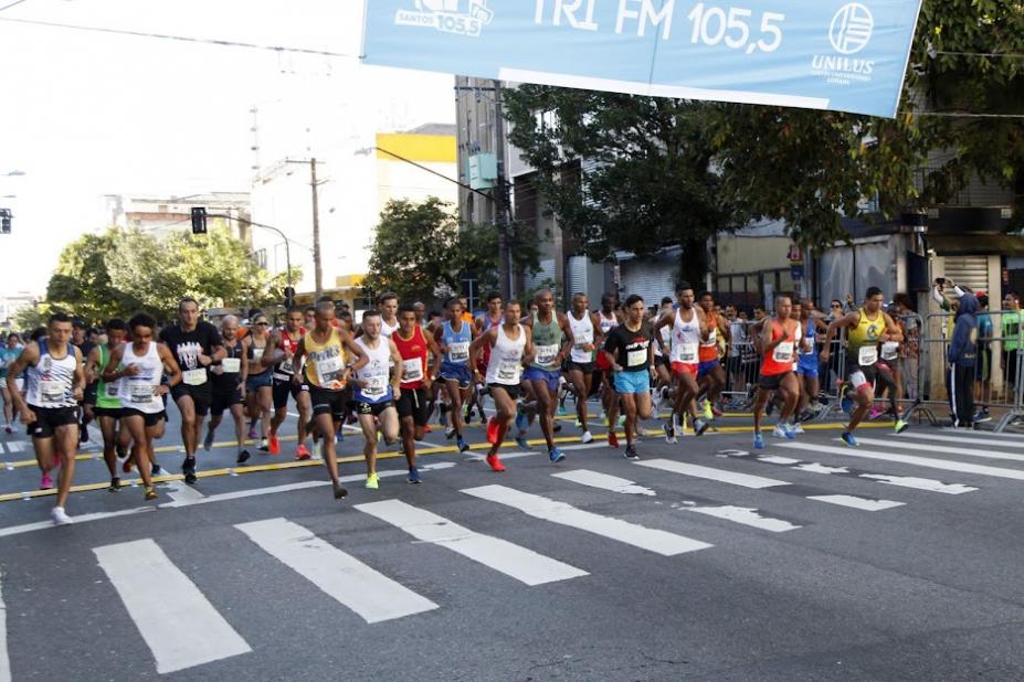 pelotão de atletas na saída de competição. Eles estão atrás de faixa de pedestres na rua. Acima, uma faixa com dizeres da prova. #paratodosverem