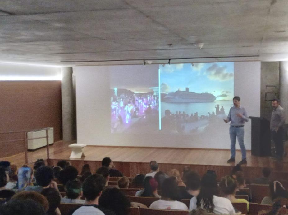 pessoas estão sentadas em auditório assistindo homem que fala diante de uma projeção. #paratodosverem