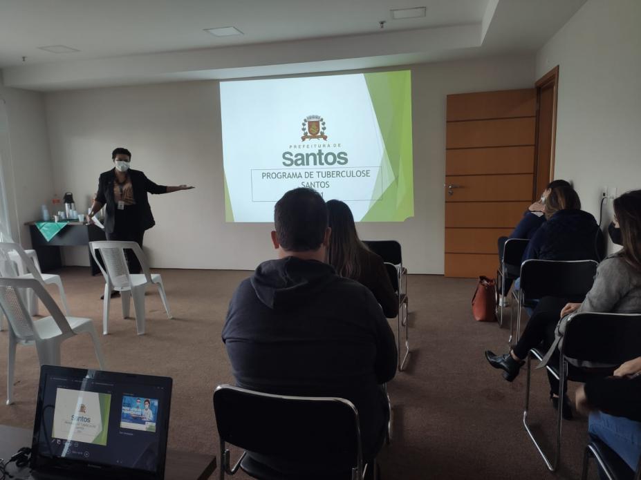 Em um auditório, mulher realiza palestra com várias pessoas assistindo. #paratodosverem