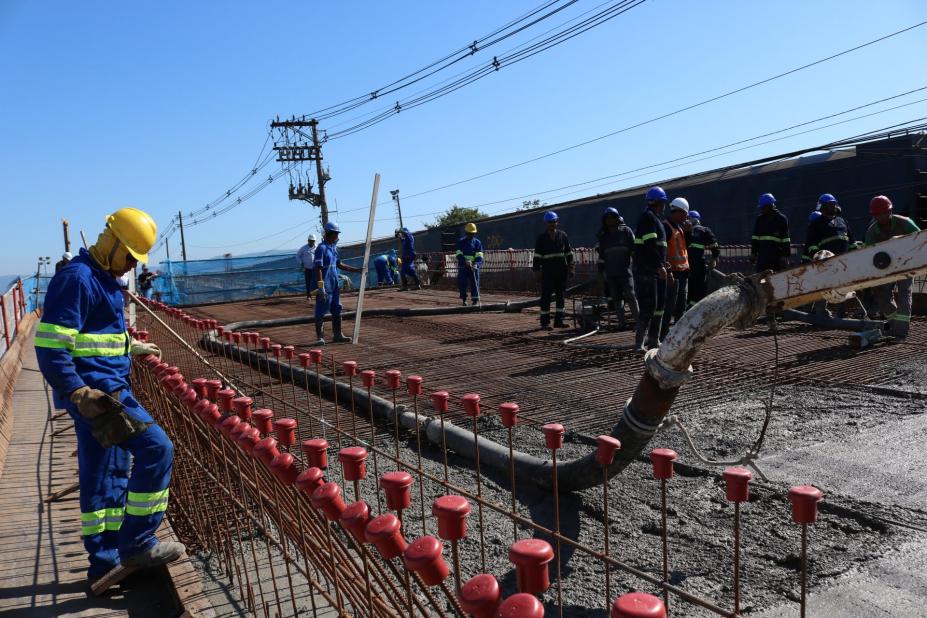 Homem com uniforme e capacete trabalha em primeiro plano enquanto outros dez aparecem ao fundo sobre pista de viaduto