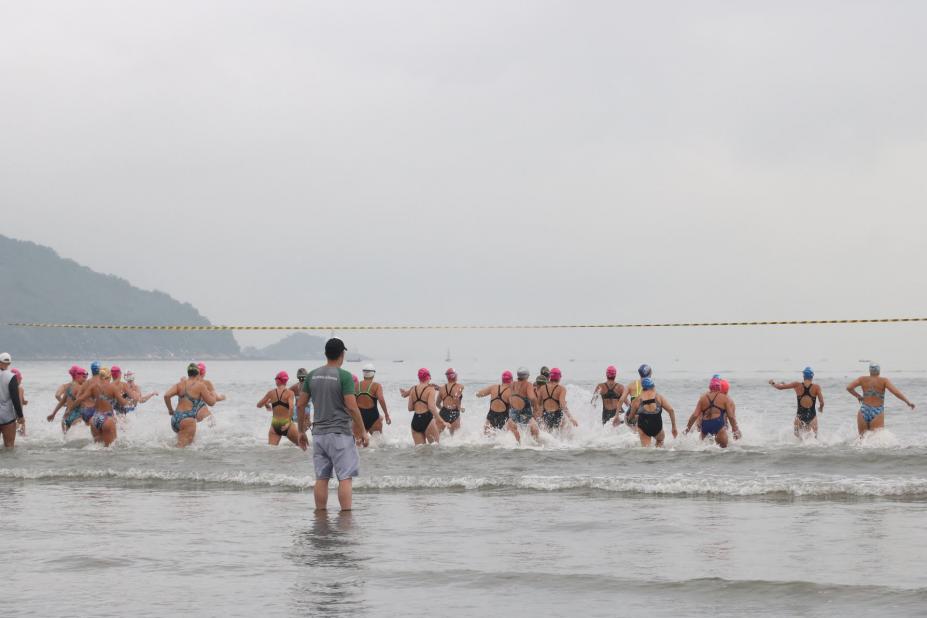 Quarenta mulheres com uniforme de natação entram no mar observadas por homem da organização