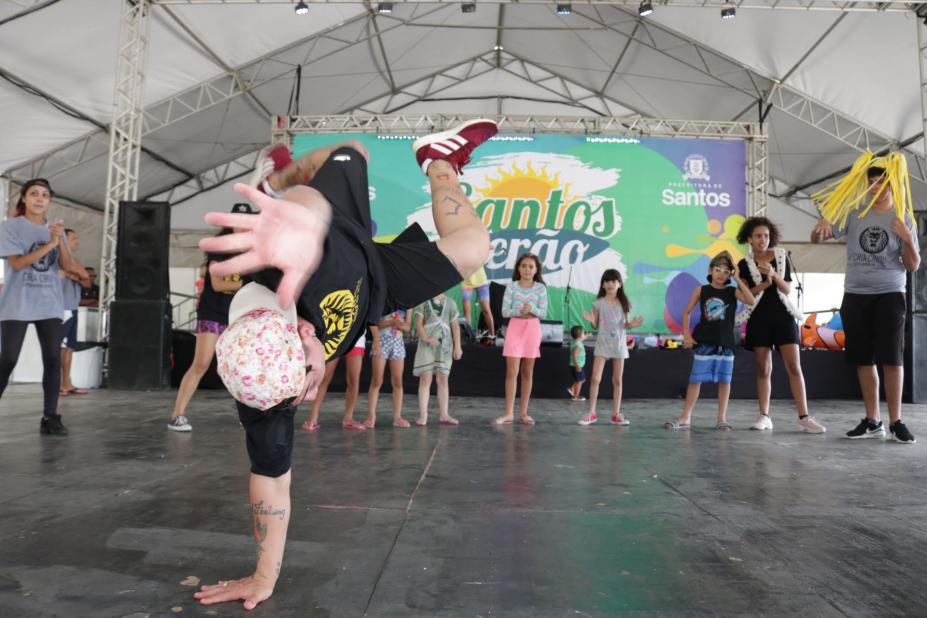 Em primeiro plano, menino faz acrobacia no chão. Atrás, onze crianças asssitem batendo palmas