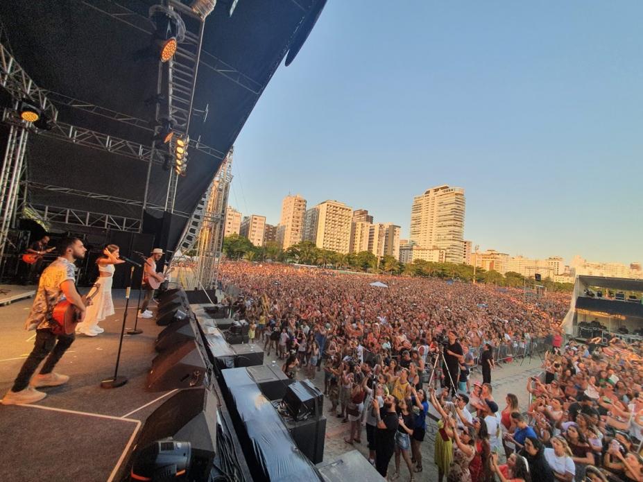 Músicos no palco e imensa multidão no chão, com vista a partir da praia para dentro da cidade. #Pracegover