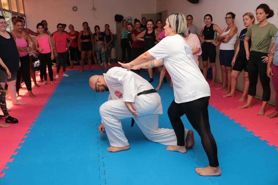 aula de defesa pessoal, mulher empurra homem pelas costas. Ele está com um dos joelhos no chão. Alunas em pé e em torno assistem à demonstração. #paratodosverem 