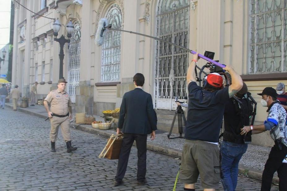 gravação de filme no centro histórico. Homem fardado caminha sobre paralelepípedo. Câmeras e microfones à frente. #paratodosverem 