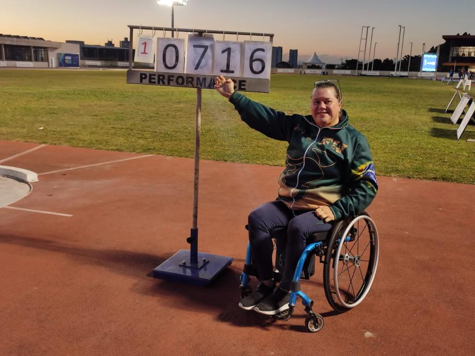 paratleta está na cadeira de rodas apontando a mão direita para o placar ao seu lado, com o número do recorde. #paratodosverem 