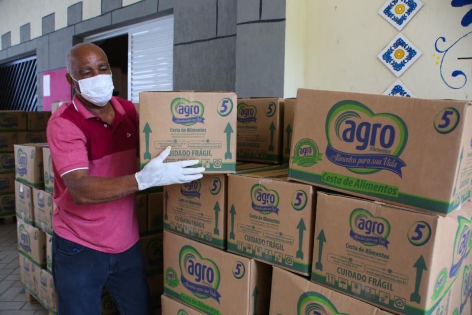 Homem ergue caixa com alimentos que está sobre uma pilha de outras caixas. O homem usa máscara e luva. #Paratodosverem