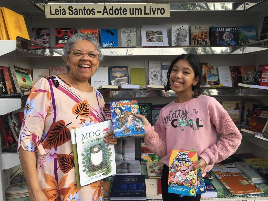 mulher e menina posam para foto segurando livros e gibis. Ao fundo, uma prateleira cheia de livros. #paratodosverem 