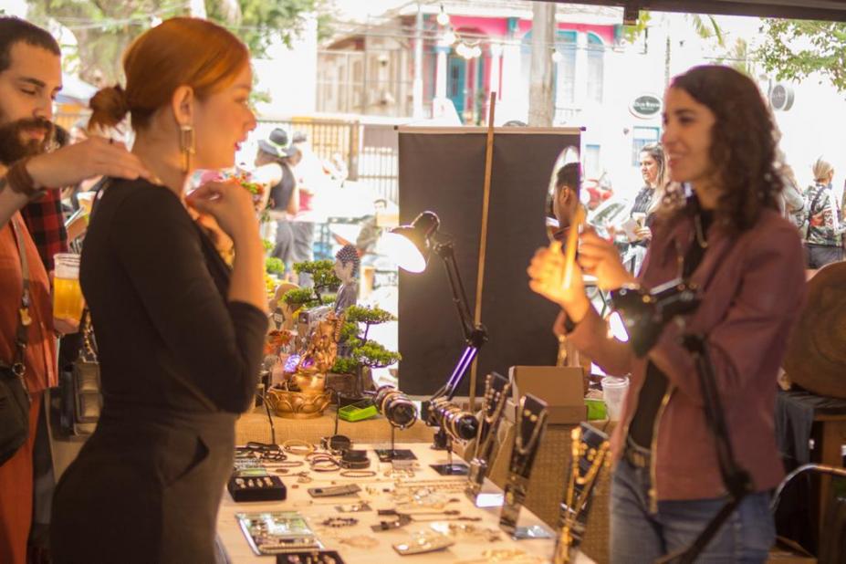 mulher experimenta colar no pescoço enquanto outra, atrás de uma mesa cheia de bijuterias, segura um espelho. #paratodosverem