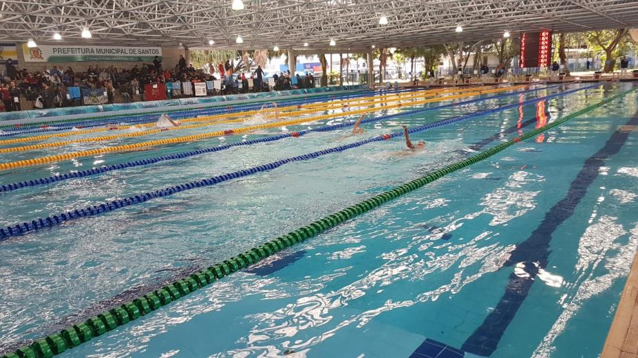 Vista geral da piscina olímpica com nadadores competindo e público ao fundo. #Pracegover