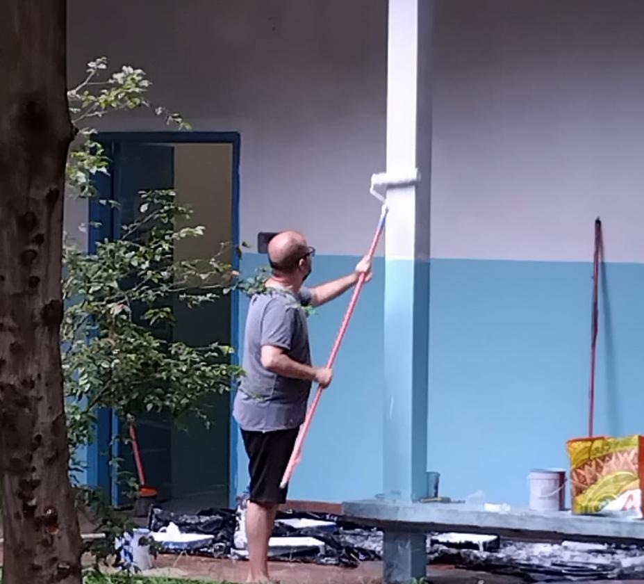 homem segura rodo de pintura com cabo longo e pinta coluna externa de escola. #paratodosverem 