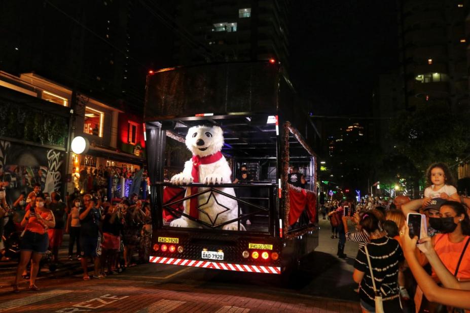 boneco de urso polar desfila em carro alegórico. Público está na rua assistindo. #paratodosverem