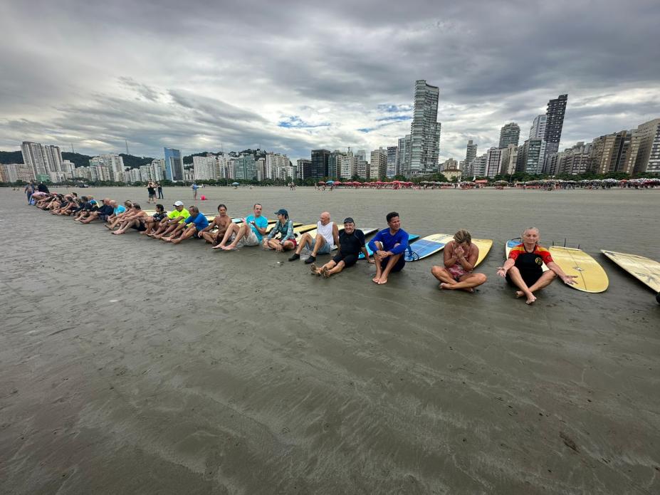 alunos estão sentados na areia em aquecimento para a aula. todos estão com pranchas. #paratodosverem