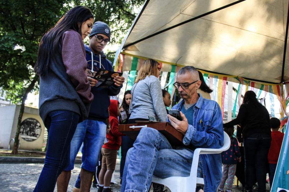 O cartunista desenha caricatura sentado em uma cadeira na praça. Há dois jovens na frente dele. #Pracegover