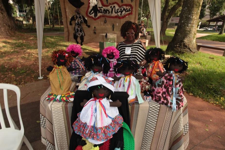 Mulher negra está à frente de banca com bonecas negras artesanais. #paratodosverem