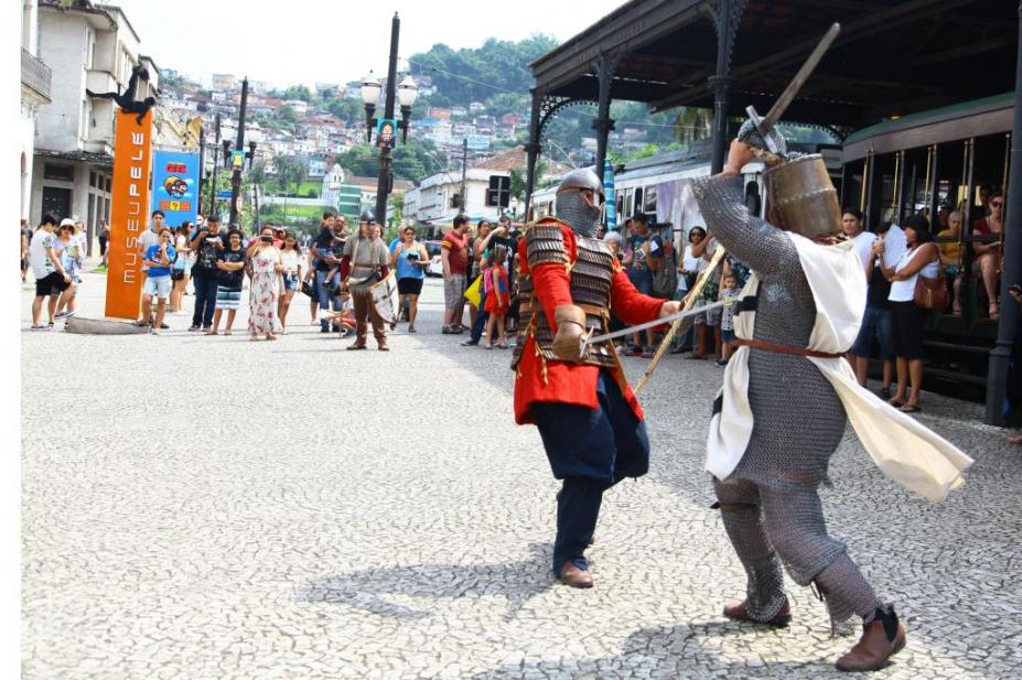 Duas pessoas fantasiadas lutam com espadas e várias pessoas assistem ao redor #paratodosverem