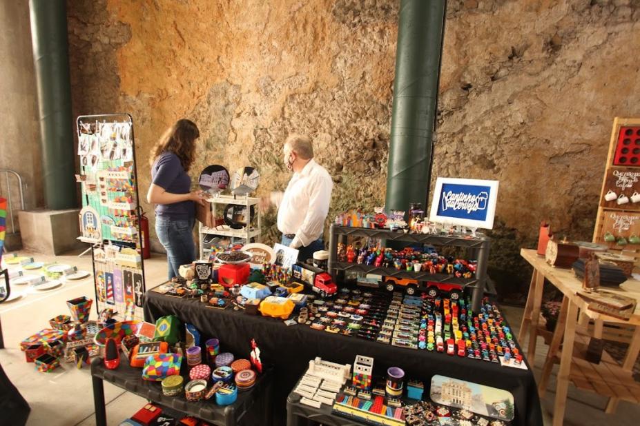 feira de artesanato com mesas cheias de pequenas peças. Duas pessoas estão em pé em torno dessas mesas. #paratodosverem 