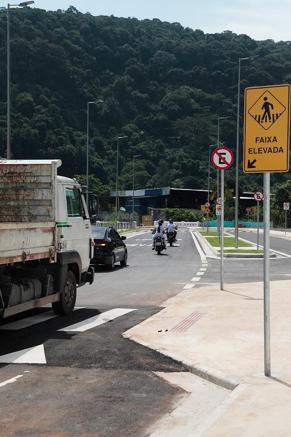 Caminhão passa por via em trecho de curva. Ele passa sobre faixa elevada. Ao lado direito, na calçada a um poste informativo sobre o elevado. Ao fundo se vê a vegetação de um morro. #Paratodosverem