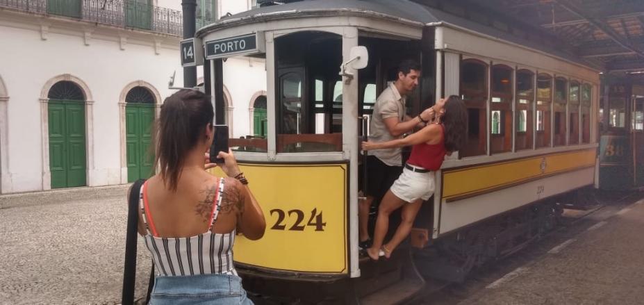 Um casal está nos degraus de entrada do bonde. Um está de frente para o outro. Do lado de fora uma mulher os fotografa com celular. #Pracegover