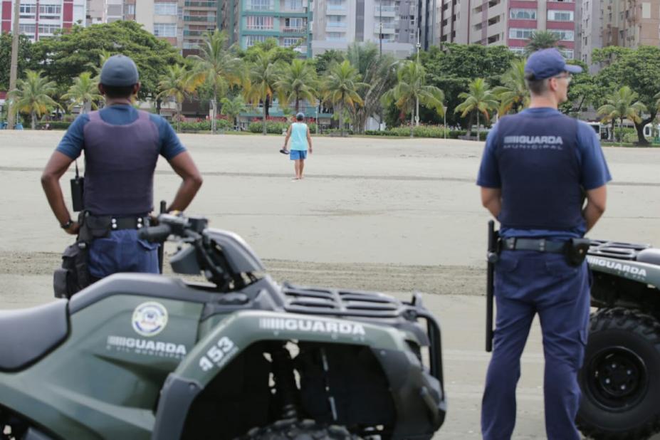 Dois guardas municipais estão na faixa de areia em pé e ao lado de quadriciclos. Á frente e distante, caminha um homem rumo ao jardim. #Paratodosverem