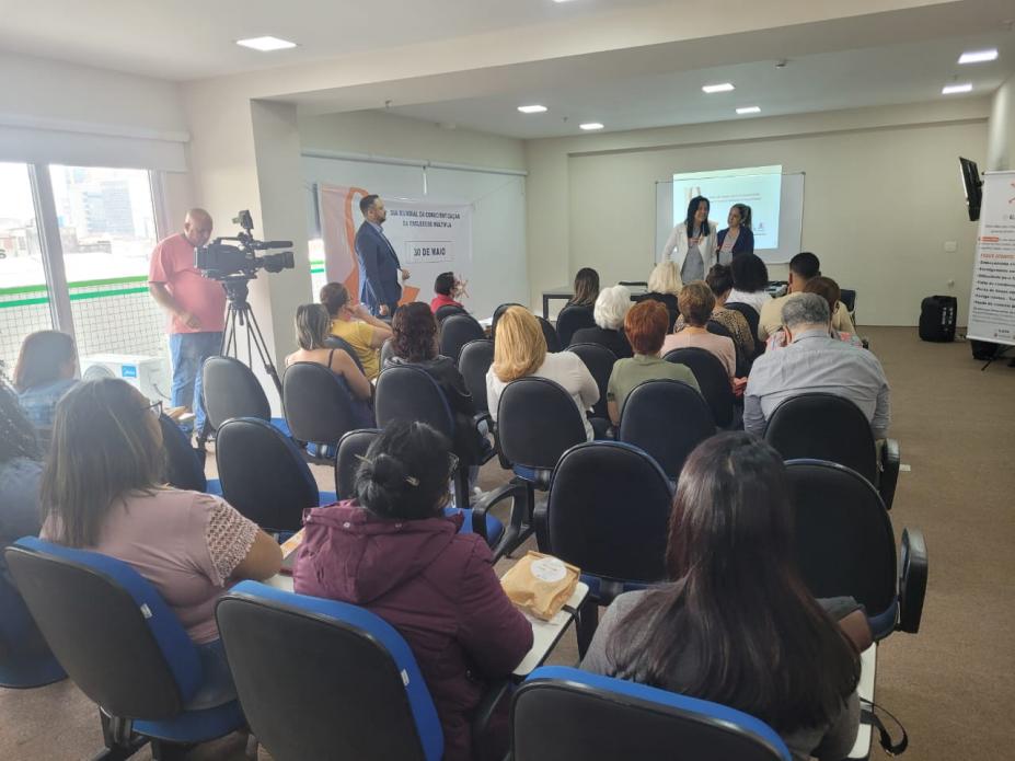 PESSOAS ESTÃO SENTADAS EM AUDITÓRIO E OUTRAS TRÊS ESTÃO EM PÉ AO FUNDO, SENDO QUE DUAS DELAS FALAM. #PARATODOSVEREM