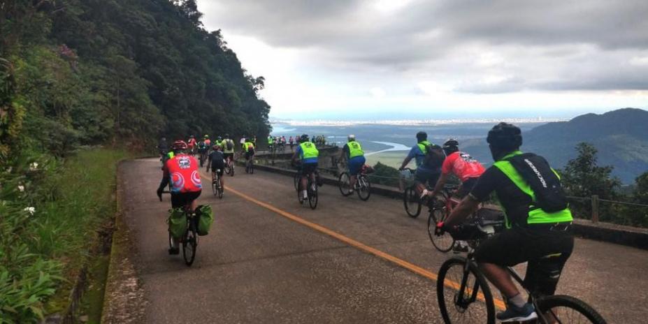 ciclistas pedalam em estrada. Vista da serra dá para a baixada santista. #paratodosverem