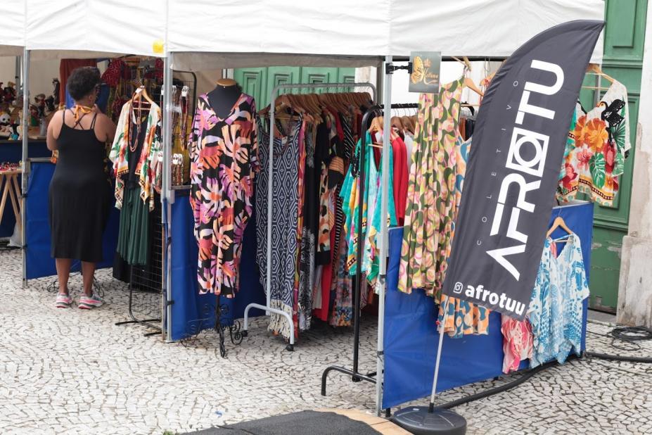 tendas sob as quais estão expostas peças de roupas. À frente de todas, um banner onde se lê afrotu. #paratodosverem 