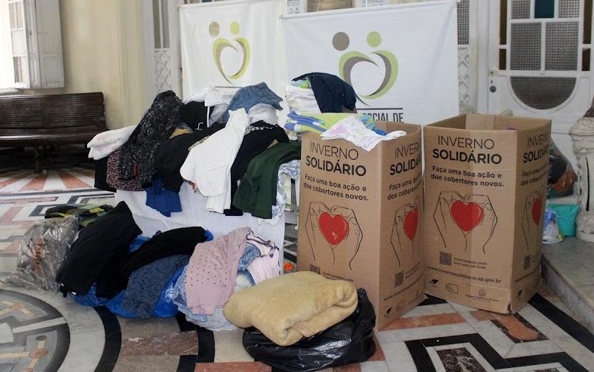 roupas empilhadas no chão e  caixas com agasalhos. Ao fundo, o banner do Fundo Social. #paratodosverem