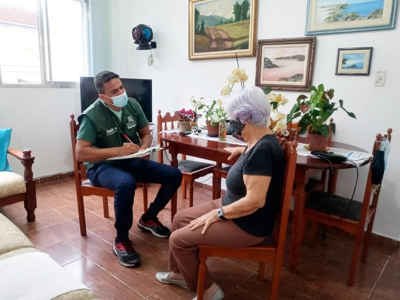 Agente comunitário, uniformizado com colete verde, conversa sentado com senhora na sua sala de jantar. #pracegover