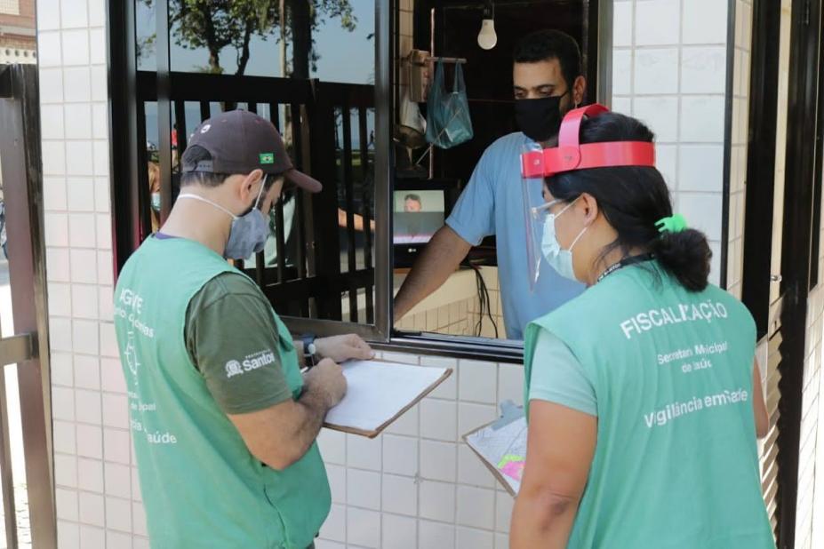 Agentes uniformizados estão em frente a guarita de prédio onde um zelador os atende. #Paratodosverem