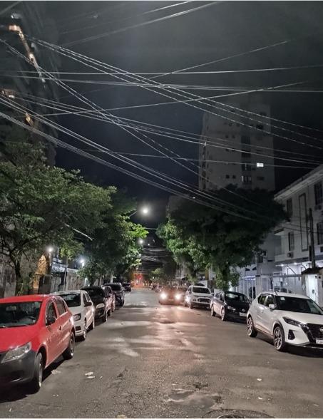rua iluminada à noite. há carros estacionados. #paratodosverem 