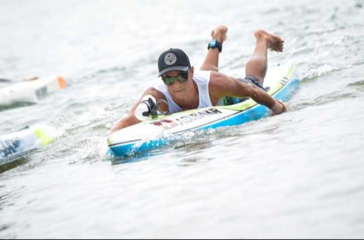 Alex está na água, deitado sobre o paddleboard. #paratodosverem