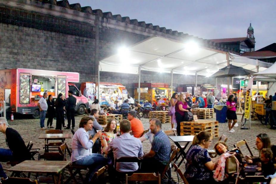 Praça de alimentação com pessoas sentadas e estandes de alimentos. #paratodosverem