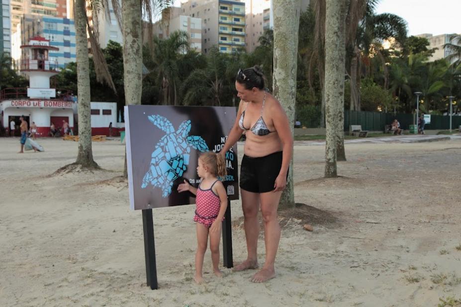 Mulher e criança observam placa educativa instalada na faixa de areia da Praia do José Menino. #pratodosverem