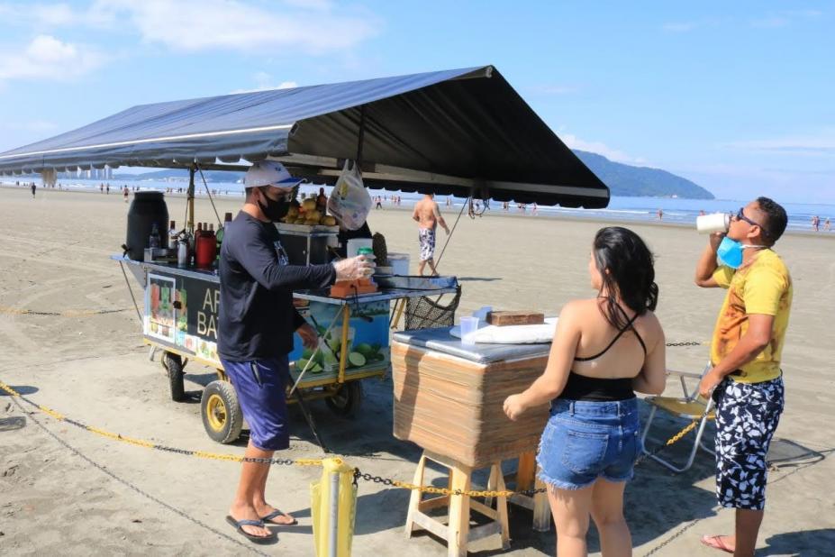 Carrinho de ambulante de bebidas cercado por faixa zebrada. O vendedor atende dois clientes na faixa de areia. #paratodosverem