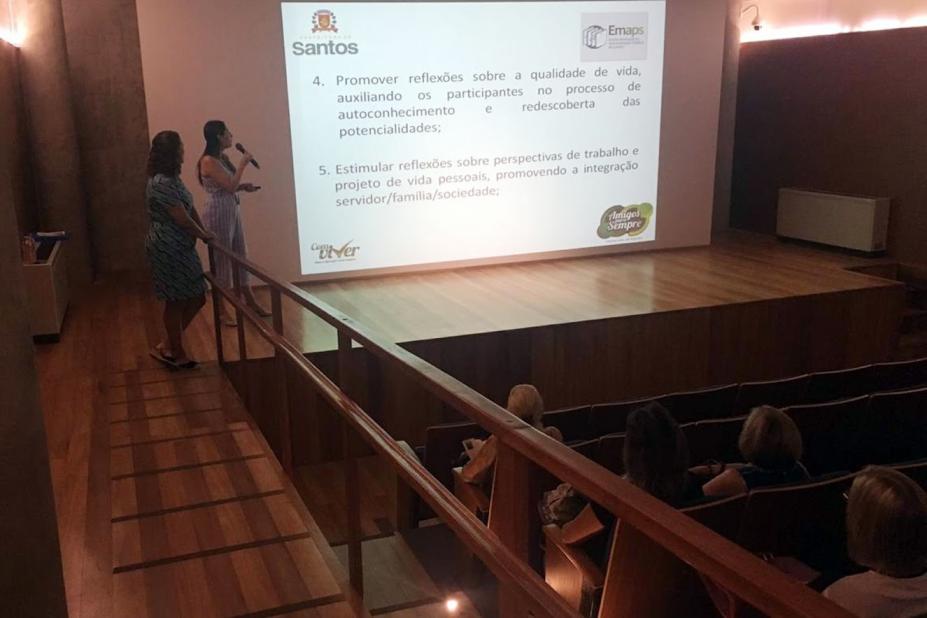 Auditório com uma mulher falando ao microfone, ao lado de imagem projetada na parede. Pessoas estão sentadas assistindo. #Paratodosverem