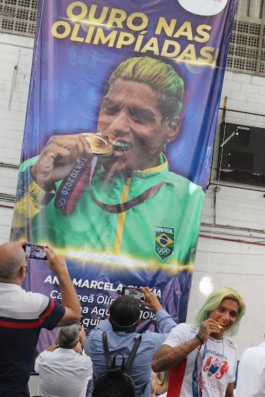 Ana Marcela exibe medalha de ouro em frente a faixa em sua homenagem. #pracegover