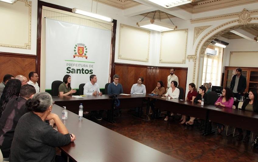 sala de reuniões com várias pessoas sentadas em uma mesa em formato de letra U. Ao fundo, o prefeito ladeado por secretários. Na parede, um banner da Prefeitura com o brasão de Santos. #paratodosverem