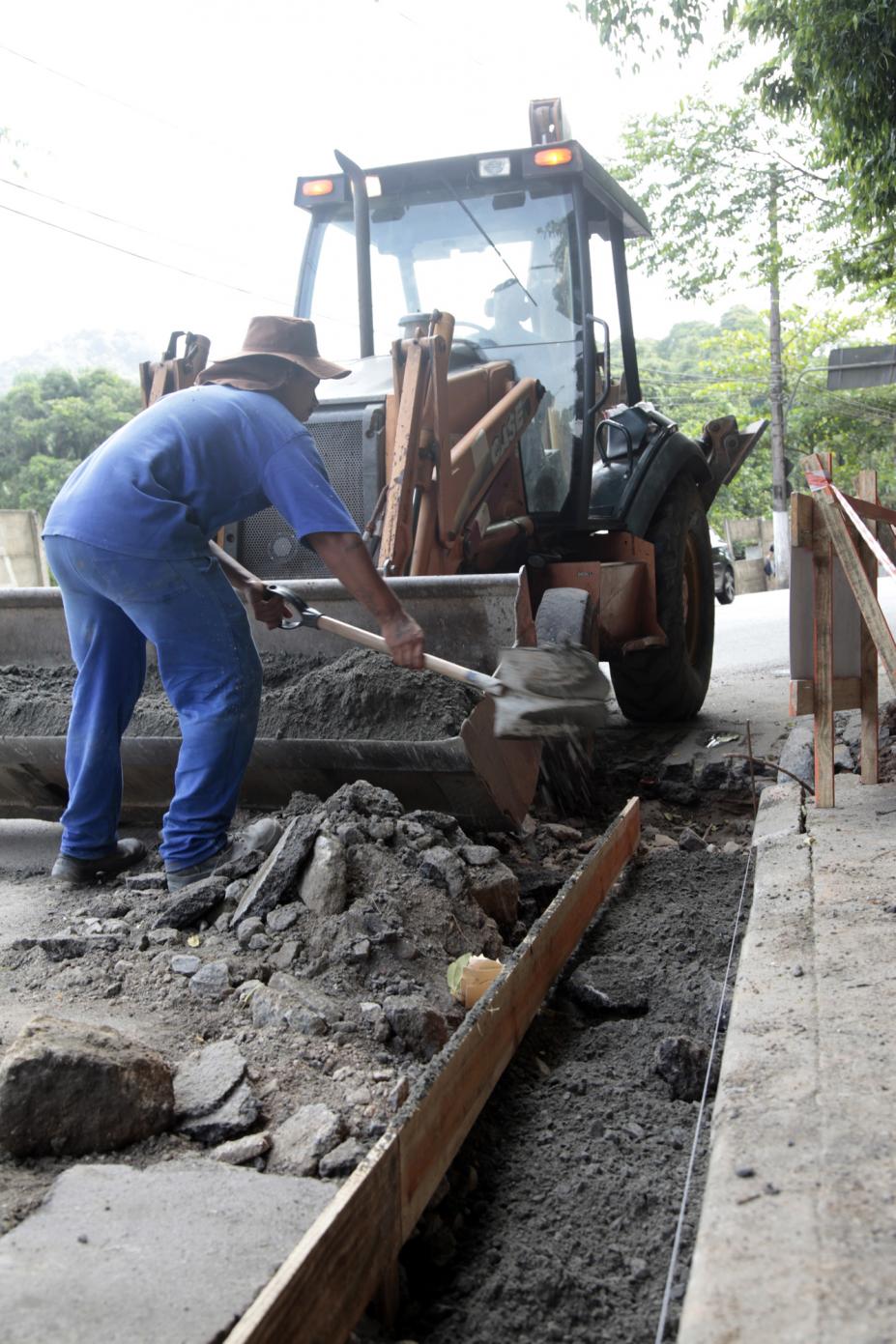 Homem está colocando cimento em via. Ele está diante de uma máquina. Ao lado, se vê a construção da sarjeta. #Pracegover