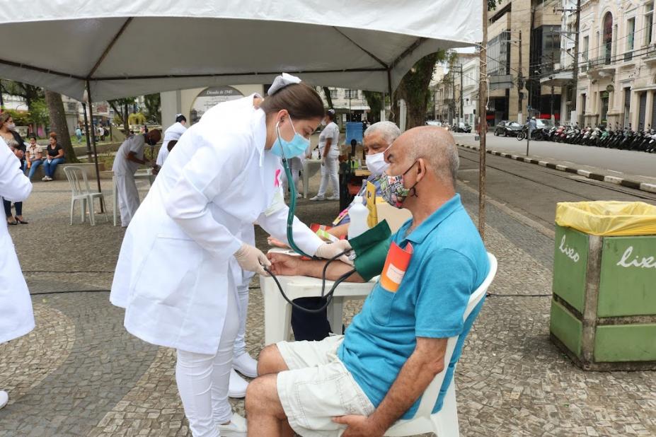 Enfermeira mede pressão de idoso sentado em cadeira na praça #paratodosverem