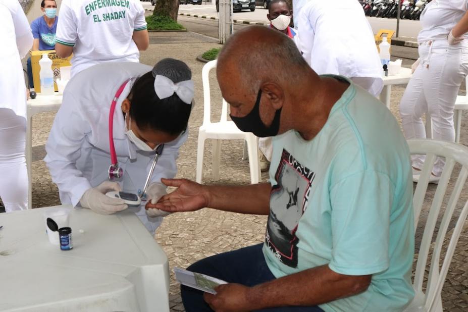 mulher vestida com avental branco e estetoscópio pendurado no pescoço aponta equipamento para medir glicemia para dedo de homem que está sentado. #paratodosverem