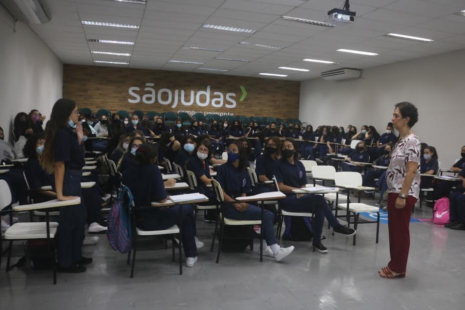 sala cheia de jovens. Uma delas está em pé falando a todas.A coordenadora da roda também está em pé. #paratodosverem