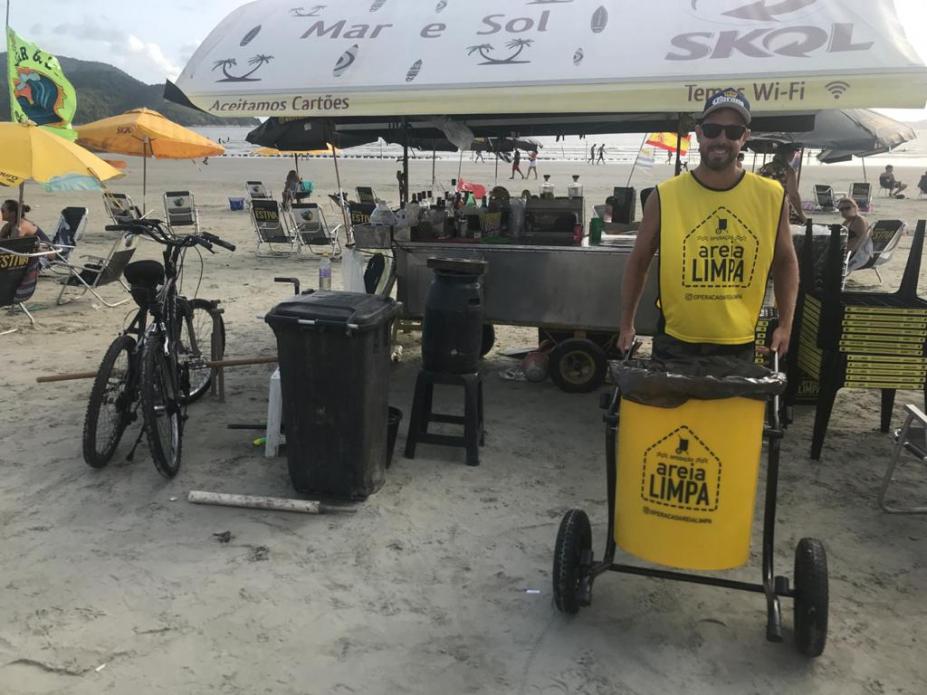 Um carrinho de bebidas está parado na areia da praia. Ele tem um toldo. Em torno dele há  uma lixeira. À frente, um homem segura um carrinho coletor de lixo pequeno. No carrinho e na camiseta do rapaz se lê: Areia Limpa. #Pracegover