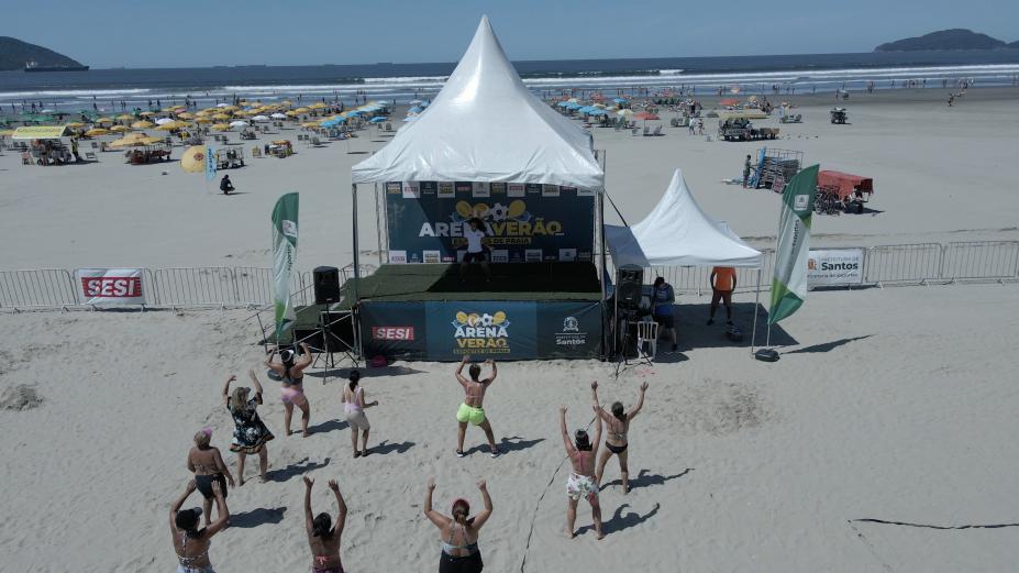 pessoas estão dançando na faixa de areia, diante de uma tenda armada. O mar está ao fundo. #paratodosverem